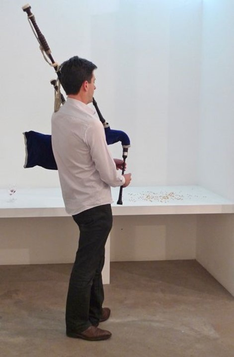 A man playing the bagpipes in a gallery space at a shelf of un-popped popcorn kernals